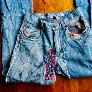 May include: Light blue denim jeans with distressed details, patches, and decorative stitching. Features a patchwork design on the front and frayed edges. The jeans are laid flat on a wooden surface.