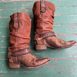 Peut inclure: Une paire de bottes de cow-boy en cuir marron avec des découpes complexes et un motif en peau de serpent sur la pointe. Les bottes ont un aspect ample et sont usées.