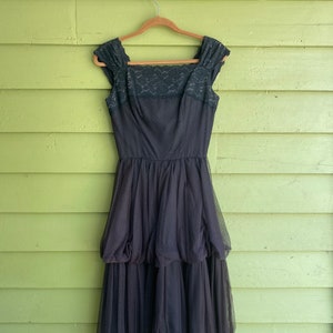 May include: A vintage black dress with a square neckline and a layered tulle skirt. The bodice features a lace overlay, and the dress hangs from a wooden hanger against a green background. A classic formal wear piece.