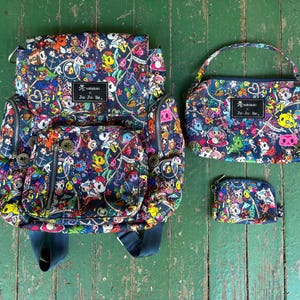 Könnte beinhalten: Ein Set von Ju-Ju-Be-Taschen mit einem farbenfrohen Cartoon-Muster. Das Set umfasst einen Rucksack, eine Handtasche und eine kleine Geldbörse. Jede Tasche hat einen dunkelblauen Hintergrund mit verschiedenen Cartoon-Figuren und dem Ju-Ju-Be-Logo.