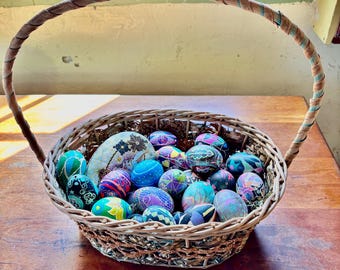 Può includere: Un cesto di vimini pieno di uova di Pasqua colorate e dipinte a mano. Le uova sono decorate con motivi e disegni intricati in tonalità di blu, verde, viola e nero.