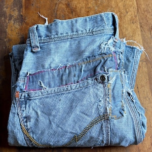 May include: Folded light blue denim jeans with distressed details. Features visible stitching in yellow, orange, and pink. The jeans have a button closure and classic five-pocket design. The fabric appears worn and faded.