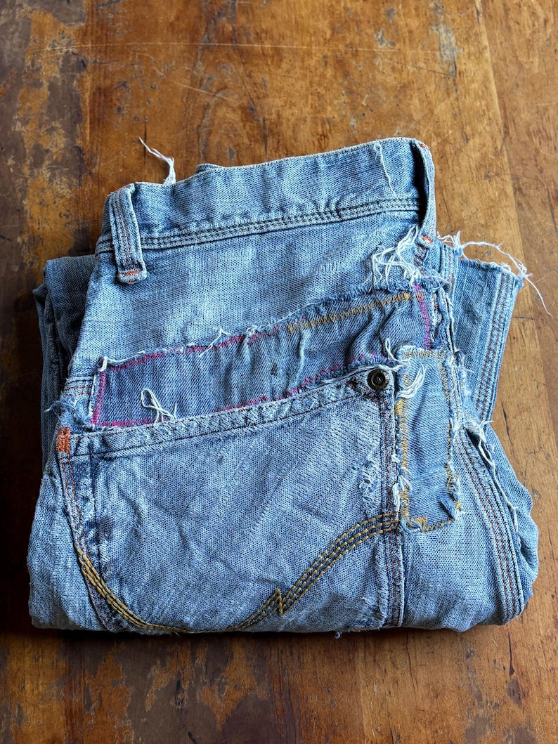 May include: Folded light blue denim jeans with distressed details. Features visible stitching in yellow, orange, and pink. The jeans have a button closure and classic five-pocket design. The fabric appears worn and faded.