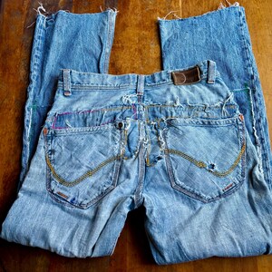 May include: Light blue denim jeans with distressed details. Features include a brown leather patch, decorative stitching, and frayed edges. The jeans are folded on a wooden surface.