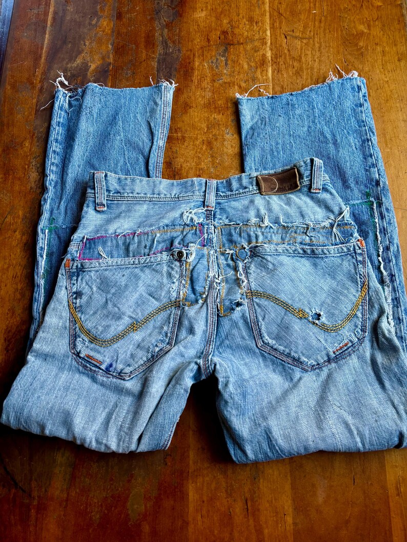 May include: Light blue denim jeans with distressed details. Features include a brown leather patch, decorative stitching, and frayed edges. The jeans are folded on a wooden surface.