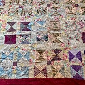 May include: A patchwork quilt with a variety of patterned squares in shades of blue, purple, pink, and brown. The quilt features floral and geometric designs, with a deep red border.