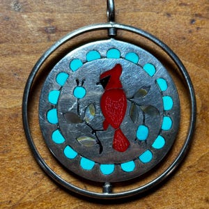 May include: A circular silver pendant featuring a rotating center disc. The disc displays a red cardinal bird perched on a branch with iridescent shell leaves, surrounded by a border of small, round, bright turquoise stone inlays.