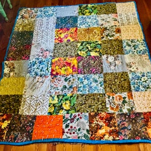 May include: A rectangular patchwork quilt constructed from various fabric squares featuring diverse floral, botanical, and abstract patterns in shades of green, orange, yellow, blue, and brown, finished with a bright blue fabric border trim.