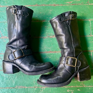 May include: A pair of black leather mid-calf boots featuring a rounded toe, a stacked block heel, and two adjustable straps with brass-toned metal buckles. One buckle is positioned across the ankle, while the other is located at the top shaft.