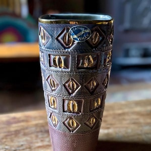 May include: A decorative vase with a unique design. The vase features a dark brown base and a textured body with repeating gold-colored geometric shapes. The rim is trimmed with gold, and a blue and white label is visible.