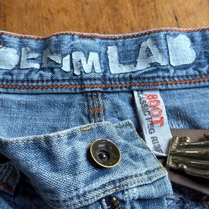 May include: Close-up of blue denim jeans with the text "DENIM LAB" across the waistband. The jeans have a button closure, a zipper, and a brown leather belt with a gold-colored buckle. The tag reads "BOOT CLASSIC PEG RISE".