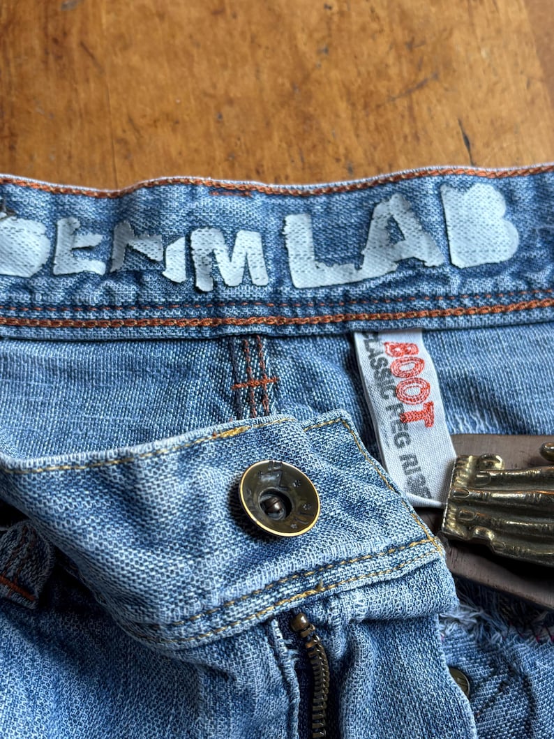 May include: Close-up of blue denim jeans with the text "DENIM LAB" across the waistband. The jeans have a button closure, a zipper, and a brown leather belt with a gold-colored buckle. The tag reads "BOOT CLASSIC PEG RISE".