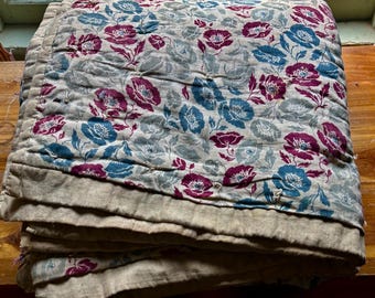Può includere: Trapunta floreale piegata con un motivo di fiori blu e bordeaux su sfondo grigio. La trapunta ha un bordo beige ed è impilata su una superficie di legno vicino a una finestra.