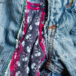 May include: Close-up of a denim garment with visible stitching and patches. A dark gray fabric with white skull patterns is sewn into the garment, framed by bright pink stitching. Green and orange stitching adds further detail.