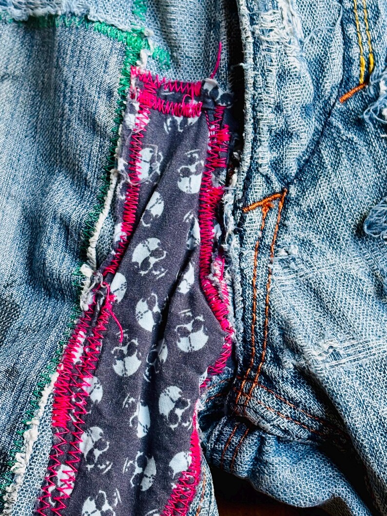 May include: Close-up of a denim garment with visible stitching and patches. A dark gray fabric with white skull patterns is sewn into the garment, framed by bright pink stitching. Green and orange stitching adds further detail.