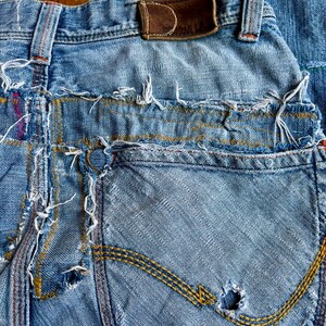 May include: Close-up of distressed blue denim jeans. The jeans feature a worn leather patch, frayed edges, and a pocket with yellow stitching. The denim has a faded appearance with visible wear and tear, including a hole in the pocket.