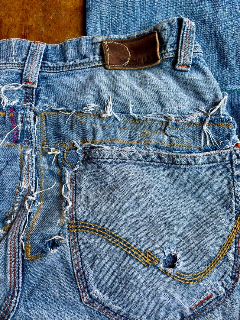 May include: Close-up of distressed blue denim jeans. The jeans feature a worn leather patch, frayed edges, and a pocket with yellow stitching. The denim has a faded appearance with visible wear and tear, including a hole in the pocket.