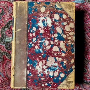 May include: An antique book with a marbled cover in shades of blue, red, and white. The spine is brown, and the corners are worn. The book is open, revealing the marbled design on the inside cover. The book is on a patterned fabric.