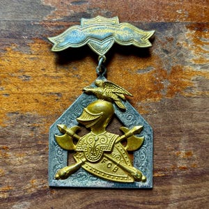 May include: Vintage metal pin with a shield design. The pin features a gold helmet, crossed axes, and an eagle. The pin has the letters "FOB" and is in shades of gold, silver, and blue.