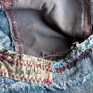 May include: Close-up of denim jeans with a visible pocket interior. The pocket lining is gray, and the seams are stitched with red thread. A label with text is sewn into the jeans with red stitching. The denim is a light blue wash.