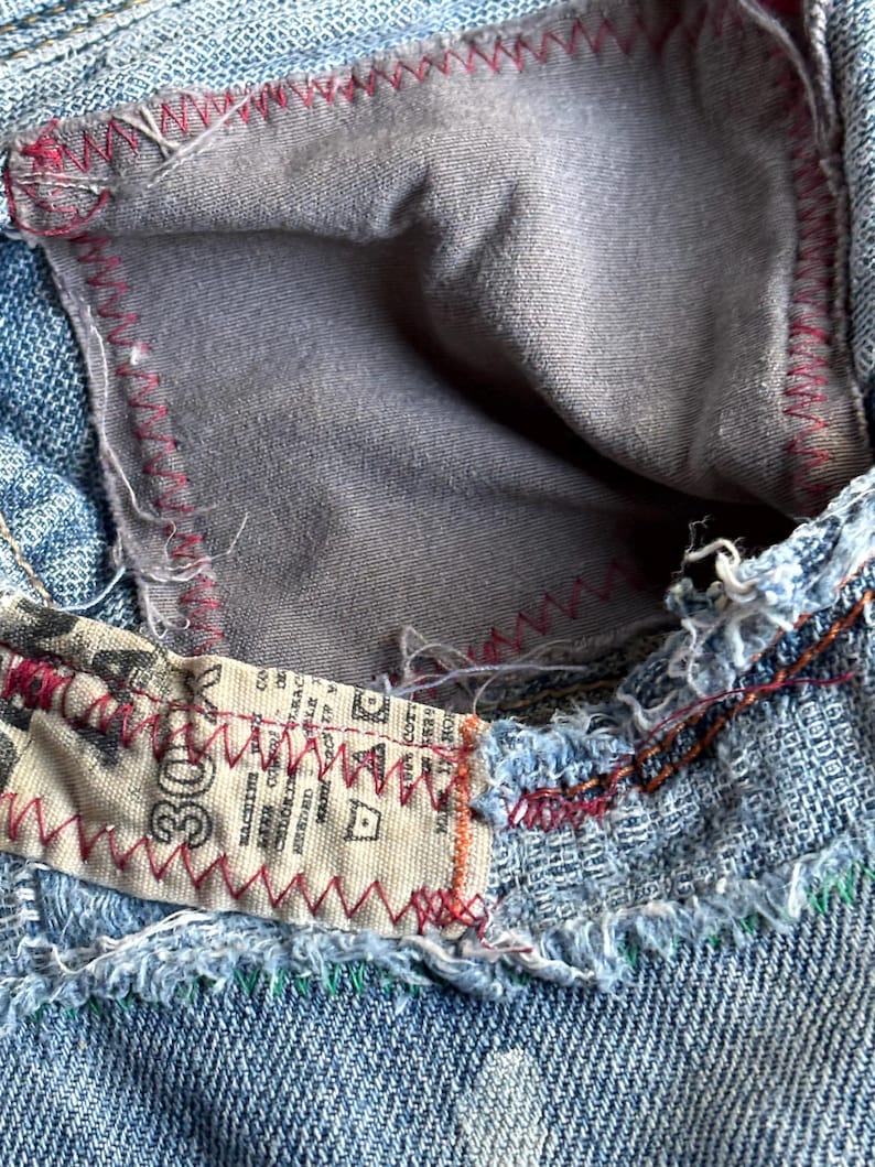 May include: Close-up of denim jeans with a visible pocket interior. The pocket lining is gray, and the seams are stitched with red thread. A label with text is sewn into the jeans with red stitching. The denim is a light blue wash.