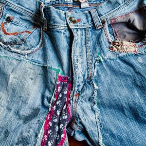 May include: Light blue denim jeans with patchwork and distressed details. Features include a button closure, decorative stitching, and a skull print fabric accent. The waistband reads "DENIM LAB".