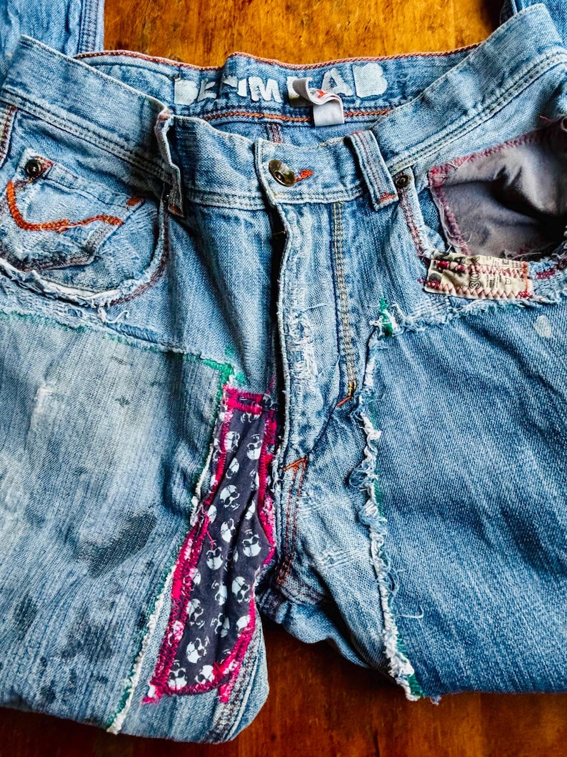 May include: Light blue denim jeans with patchwork and distressed details. Features include a button closure, decorative stitching, and a skull print fabric accent. The waistband reads "DENIM LAB".