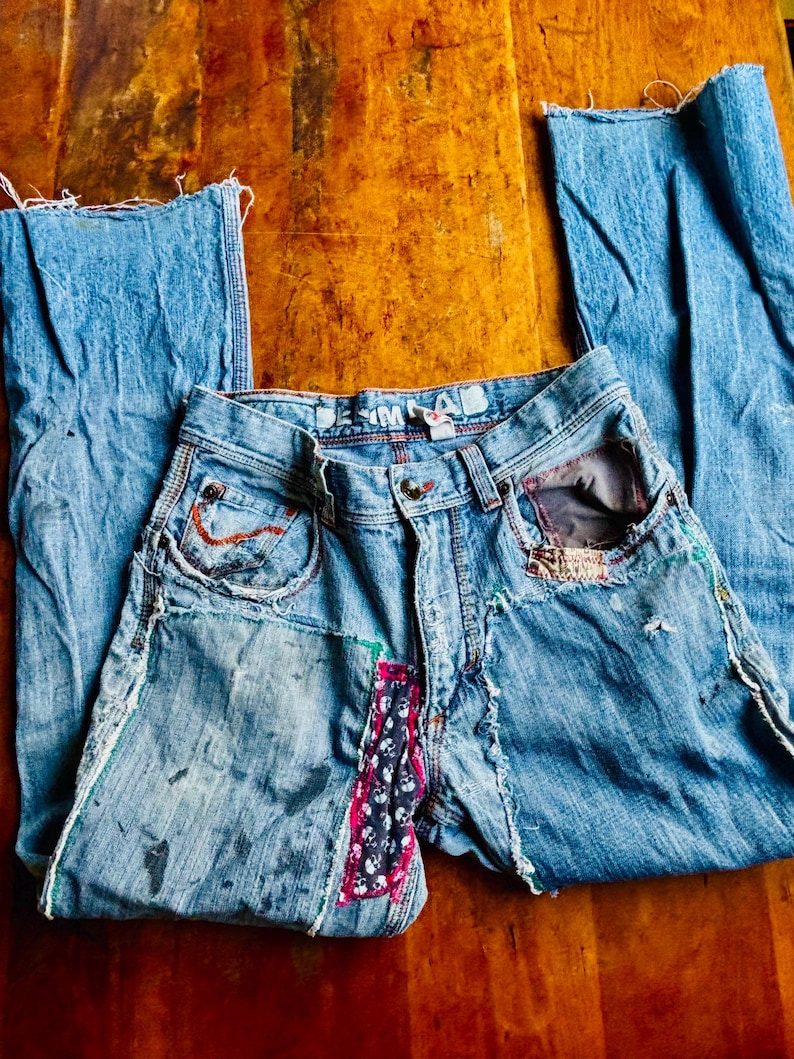 May include: Light blue denim jeans with distressed details, patches, and decorative stitching. Features a patchwork design on the front and frayed edges. The jeans are laid flat on a wooden surface.