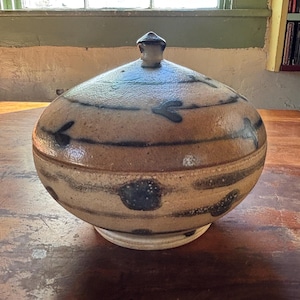 May include: A round, lidded stoneware jar with a light brown base and dark blue decorative stripes and leaf-like patterns. The jar sits on a white base and has a small knob on the lid.