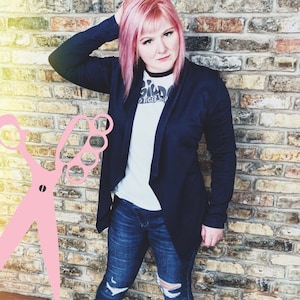 Op de afbeelding: Een vrouw met roze haar draagt een donkerblauwe cardigan over een wit T-shirt met de tekst "Old Tiger" erop gedrukt. Ze draagt ook blauwe jeans met distressed details.