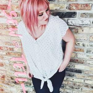 Op de afbeelding: Een vrouw met roze haar draagt een wit bloesje met een klein bloemetjespatroon. Het bloesje heeft een geknoopte voorkant en de vrouw draagt een zwarte broek.
