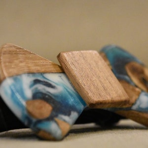 May include: A wooden bow tie with a blue and white resin inlay. The bow tie has a natural wood finish and a black adjustable strap.