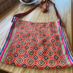 May include: A colorful, handmade shoulder bag with a geometric pattern. The bag features a vibrant orange background with diamond-shaped designs in pink, green, and navy blue. The edges are trimmed with stripes of pink, green, blue, and orange. The strap is a woven design.