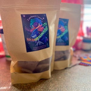 May include: Three brown paper bags of LunaAmor Cacao. Each bag has a colorful label with a dragon-like creature, a pyramid, and the text "LunaAmor Cacao". The bags are on a speckled surface.