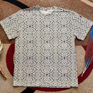 Puede incluir: Una camiseta blanca con un patrón geométrico en tonos rojos, azules, verdes y negros. El patrón es intrincado y detallado, y cubre toda la camiseta.