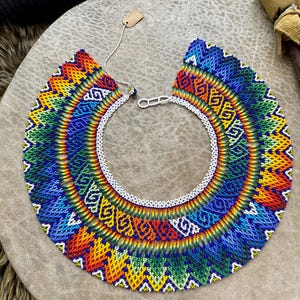Puede incluir: Un vibrante collar con cuentas en colores del arcoíris, que incluye rojo, naranja, amarillo, verde y azul. El collar presenta intrincados patrones geométricos y un collar interior de cuentas blancas. Una pequeña etiqueta cuelga del collar.