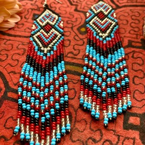 May include: A pair of long, beaded earrings with a geometric pattern in red, black, turquoise, and silver. The earrings are made of small, round beads and have a fringe-like design.