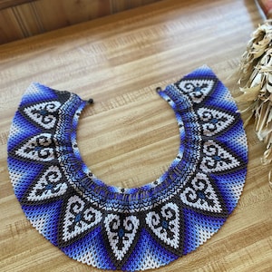 May include: A beaded collar necklace in shades of blue, black, and white. The necklace features a geometric pattern with intricate designs. The collar is displayed on a wooden surface.