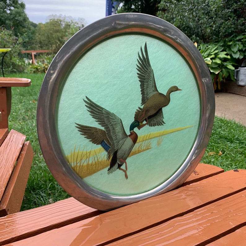 May include: Round metal tray with a painted scene of two ducks in flight over a marsh. The ducks are depicted with detailed feathers and vibrant colors. The tray has a silver-colored metal rim and a light green background.