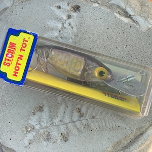 Könnte beinhalten: Ein Storm Hot'N Tot Angelköder in einer transparenten Kunststoffverpackung. Der Köder ist silber, klar und gelb mit einem gelben Auge und einer silbernen Klinge. Die Verpackung trägt die Worte "Storm Hot'N Tot" in Rot und Gelb.