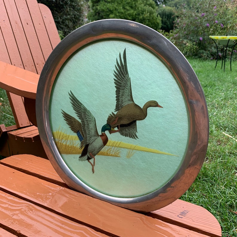 May include: Round serving tray with a silver-colored metal rim. The tray features a light green background with a detailed illustration of two ducks in flight. The ducks have brown, blue, and white feathers.