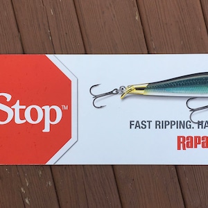 Könnte beinhalten: Ein weißes rechteckiges Schild mit einem roten Achteck mit der Aufschrift "RipStop" und einem Angelköder. Der Köder ist silber, schwarz und goldfarben mit zwei Haken. Der Text darunter lautet "FAST RIPPING. HARD STOPPING. Rapala."