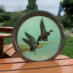 May include: Round metal tray with a painted scene of two ducks in flight over a marsh. The ducks have brown, blue, and green feathers. The tray has a silver-colored rim and a light green background.