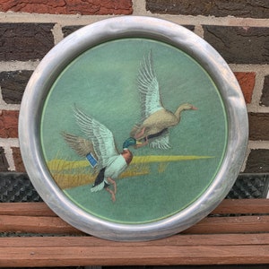 May include: Round metal tray with a painted scene of two ducks in flight over a marsh. The ducks have brown, blue, and green feathers. The tray has a silver-colored rim and a green background.