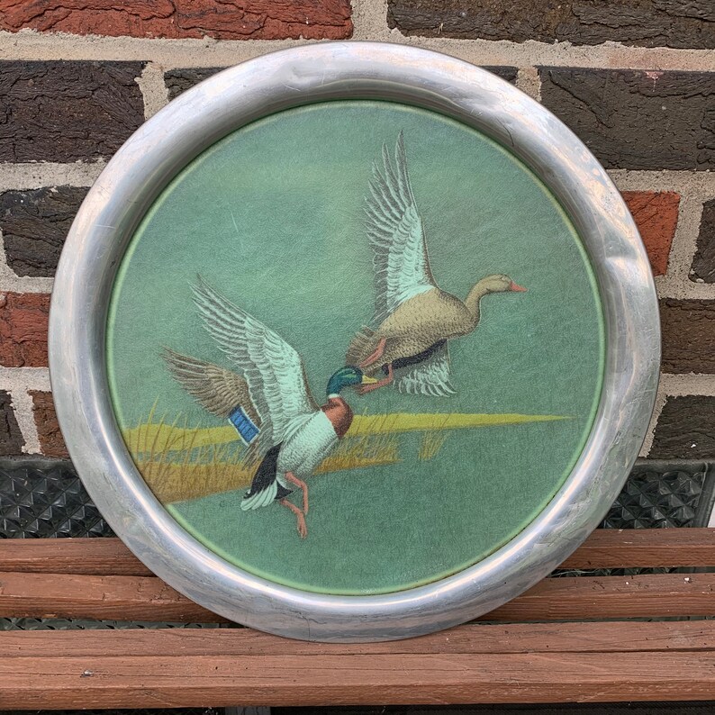 May include: Round metal tray with a painted scene of two ducks in flight over a marsh. The ducks have brown, blue, and green feathers. The tray has a silver-colored rim and a green background.