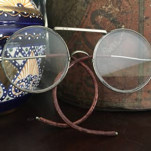 May include: Antique round eyeglasses with a silver frame and a brown leather bridge. The glasses are resting on a dark surface, with a blue and white ceramic vase and a decorative tin in the background.