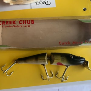 Puede incluir: Un señuelo de pesca Creek Chub vintage en su caja original. El señuelo es segmentado, con un cuerpo verde oscuro y marrón, detalles rojos y tres anzuelos triples. La caja dice "Creek Chub True-to-Nature Lures" y "Catch More Fish".