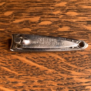 May include: A vintage chrome nail clipper with the text "'61 FORD LUBRICATES ITSELF" engraved on the surface. The clipper is lying on a wooden surface with a visible wood grain pattern.