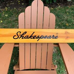 Puede incluir: Un letrero de madera con el nombre "Shakespeare" en escritura negra. El letrero es rectangular y está hecho de madera de color claro, apoyado en una silla de madera marrón. El fondo es hierba verde.