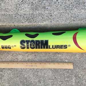 Könnte beinhalten: Ein großer, grün-gelber Angelköder mit schwarzen Flecken und dem Text "CHUG BUG" und "STORM LURES". Der Köder hat ein großes Auge und ein rotes Munddesign. Ein Holzlineal liegt neben dem Köder.